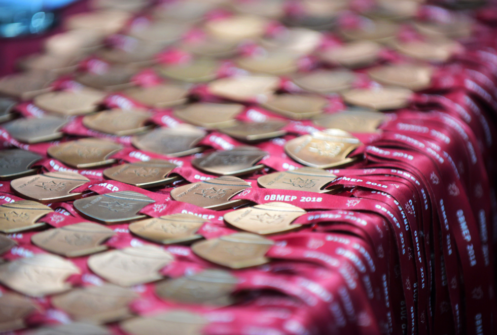 Num ambiente interno, sobre uma mesa, coberta com pano cor de vinho, seis fileiras de medalhas, sendo de prata e bronze, presas em fitas vermelhas, escrito nelas, OBMEP 2018. Imagem 8 de 13  