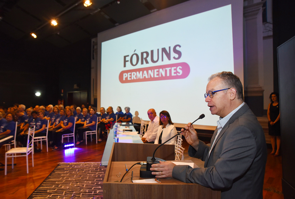 No palco de um auditório, imagem em perfil e de busto, homem em pé, à direita, fala em microfone de mesa que está sobre uma tribuna de madeira à frente dele. Ele mantém a mão direita no microfone e a esquerda na base do equipamento. Ao fundo da imagem, ampla tela de data show onde se lê Fóruns Permanentes. À esquerda na imagem, no palco, grupo com cerca de cinquenta idosos sentados em cadeiras de ferro, um ao lado do outro, formando seis fileiras. Todos usam camisetas iguais e na cor azul. Imagem 7 de 13.