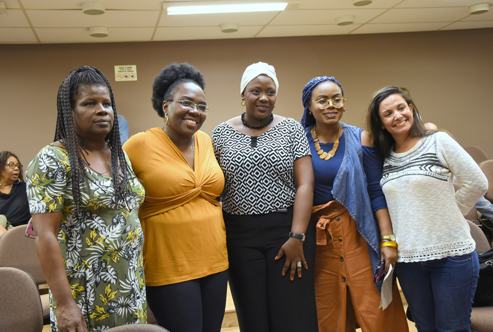 Imagem de um ambiente fechado, com cinco mulheres de pé lado a lado. Da esquerda para a direita, a primeira tem os braços ao lado do corpo, a segunda abraça as duas ao seu lado, a do centro tem o braço esquerdo à frente do corpo com a mão sobre a coxa, a quarta segura alguns papéis na mão esquerda e a quinta tem o braço flexionado em noventa graus, com a mão esquerda atrás do corpo. Atrás delas há cadeiras de auditório e ao fundo uma parede bege. Imagem 4 de 7.