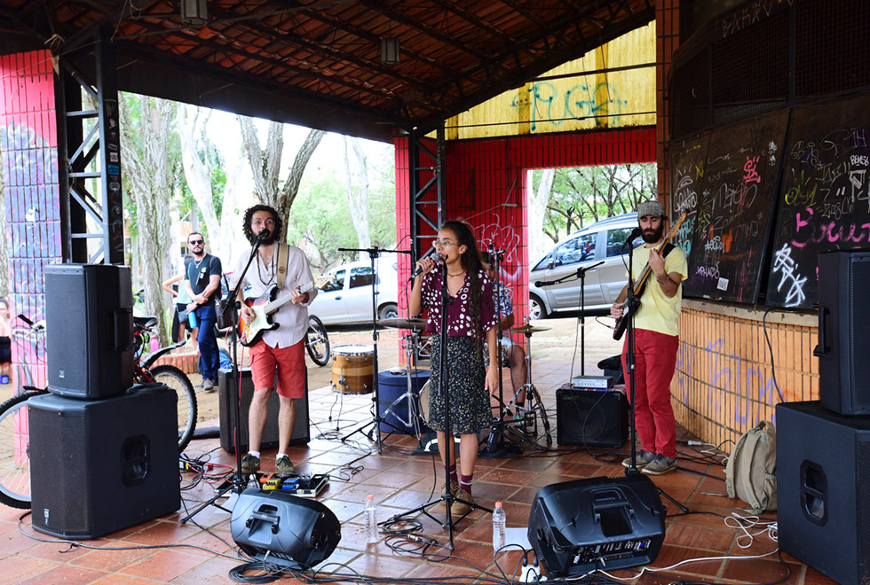 Imagem de ambiente externo. Sob uma cobertura, um grupo de três pessoas se apresenta. Dois homens tocam guitarra e uma mulher canta. As paredes e pilastras do ambiente são de tijolos-à-vista e há gravite em todas elas. Em torno dos três há microfones, caixas de som e outros equipamentos musicais. Ao fundo há um campo aberto com árvores, carros estacionados e algumas pessoas. Imagem 2 de 9.