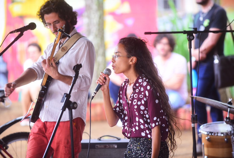 Imagem de ambiente aberto. Duas pessoas, um homem e uma mulher, estão no centro da foto se apresentando. O homem toca uma guitarra e a mulher segura um microfone, cantando. Em torno deles há diversos equipamentos musicais. Ao fundo, desfocados, pode-se ver algumas pessoas sentadas assistindo ao show, além de algumas árvores e uma parede vermelha com grafites coloridos. Imagem 3 de 9.
