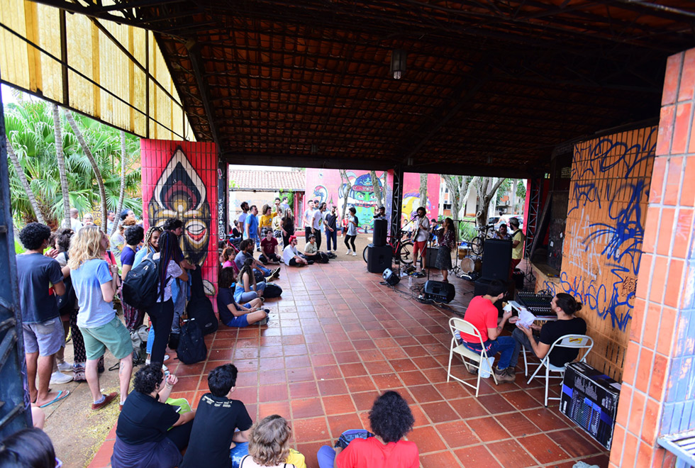 Imagem aberta de ambiente externo, tirada levemente do alto. Num espaço coberto, um grupo de jovens, alguns de pé outros sentados, assiste a uma apresentação musical ao vivo. Quatro jovens, três homens e uma mulher, ao fundo da imagem, se apresentam. Dois homens tocam guitarras e um toca bateria, enquanto a mulher canta. Há vários equipamentos de som em torno deles. Fora do espaço coberto se vêem árvores e outros prédios Á distância. Imagem 4 de 9.