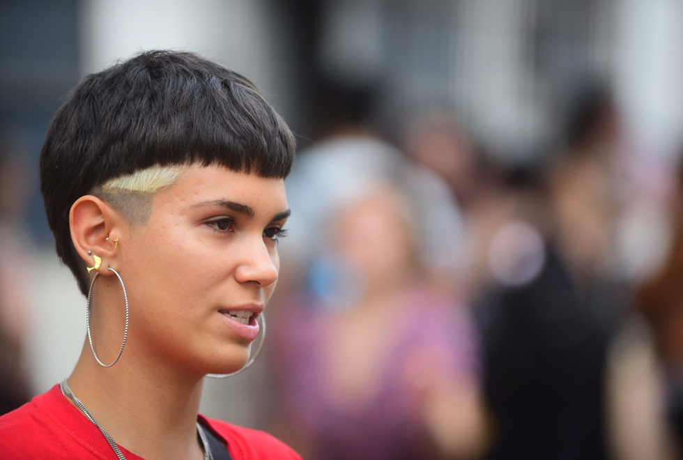 Imagem em close, de perfil, de uma mulher. Ela tem cabelos curtos e pretos com uma mecha branca, alguns piercings e usa um brinco de argola prateado. Veste uma roupa blusa vermelha e tem um colar prateado no pescoço. Ao fundo, desfocadas, podem se ver algumas pessoas de pé. Imagem 7 de 9.