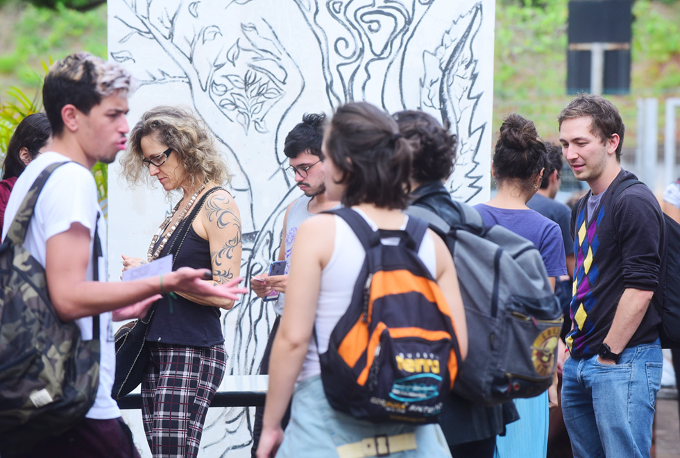 Imagem de ambiente externo. Em primeiro plano, há algumas pessoas em pé conversando. Eles se vestem casualmente e alguns carregam mochilas. Atrás deles se vê uma parede branca com uma imagem de estilizada de uma árvore desenhada em preto. Ao fundo da imagem, desfocados, há algumas árvores e paredes de um prédio mais distante. Imagem 9 de 9.