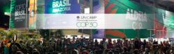 Em primeiro plano, uma grande multidão de pessoas está aglomerada, olhando para frente e para cima, em direção a um palco ou prédio. Acima da multidão, o fundo é dominado por uma série de banners verticais muito grandes, iluminados por luzes vindas de baixo. No centro exato da imagem, há uma faixa horizontal branca e semitransparente sobreposta que contém o logotipo da Unicamp e o texto "UNICAMP NA COP30". Os banners no fundo também promovem o evento. Neles, é possível ler textos como "COP30 BRASIL AMAZÔNIA BELÉM 2025" e "ALDEIA COP". À esquerda, é possível ver algumas folhas de palmeira.
